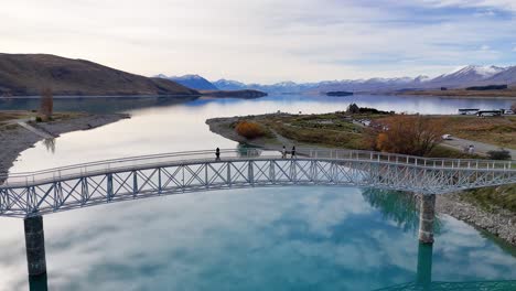 drone footage captures tourists walking across a bridge over lake tekapo, surrounded by clear blue water and mountainous landscape