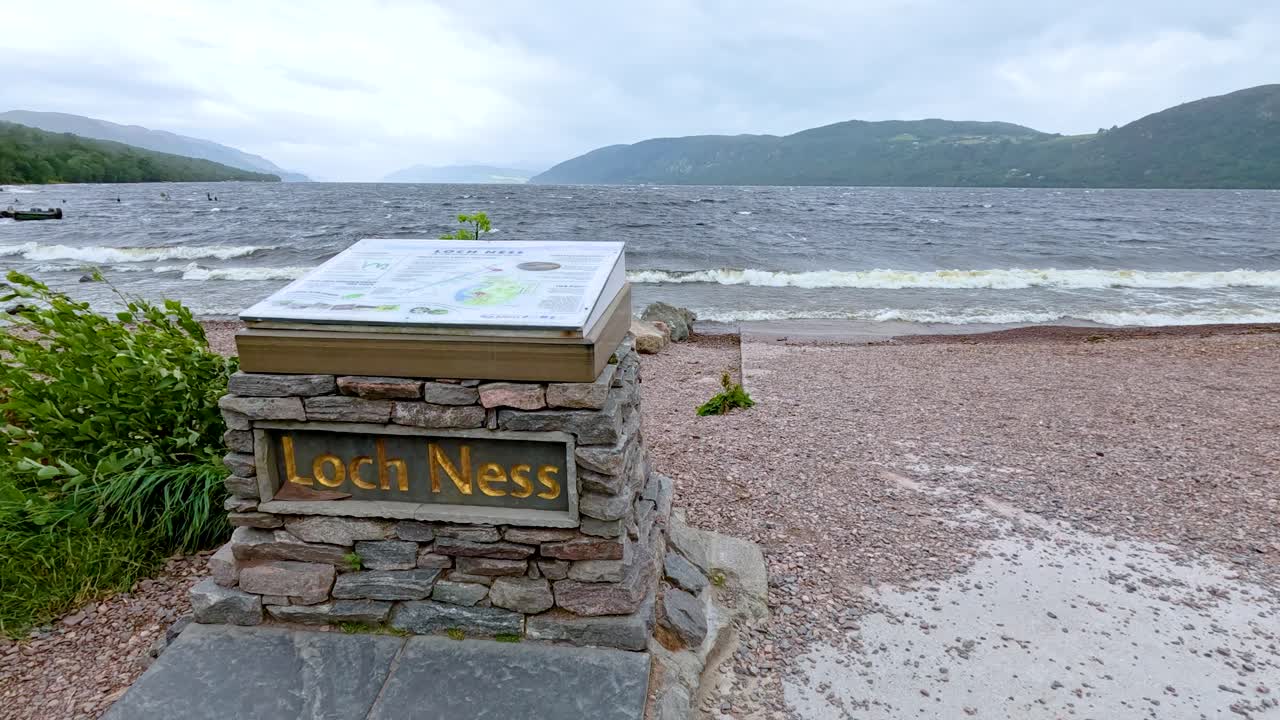 Camera approaches Loch Ness sign on pebble beach with waves, cloudy sky, and Highland hills
