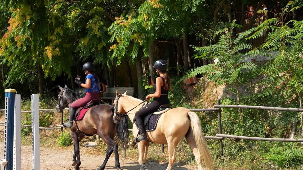 Two horse riders circle and record themselves as the coach sits in the shade reviewing the day