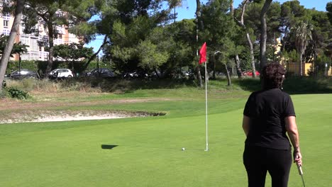 Elderly-golf-player-walks-on-a-golf-green-next-to-a-bunker-in-an-urban-field,-with-the-red-flag-and-the-put-in-hand