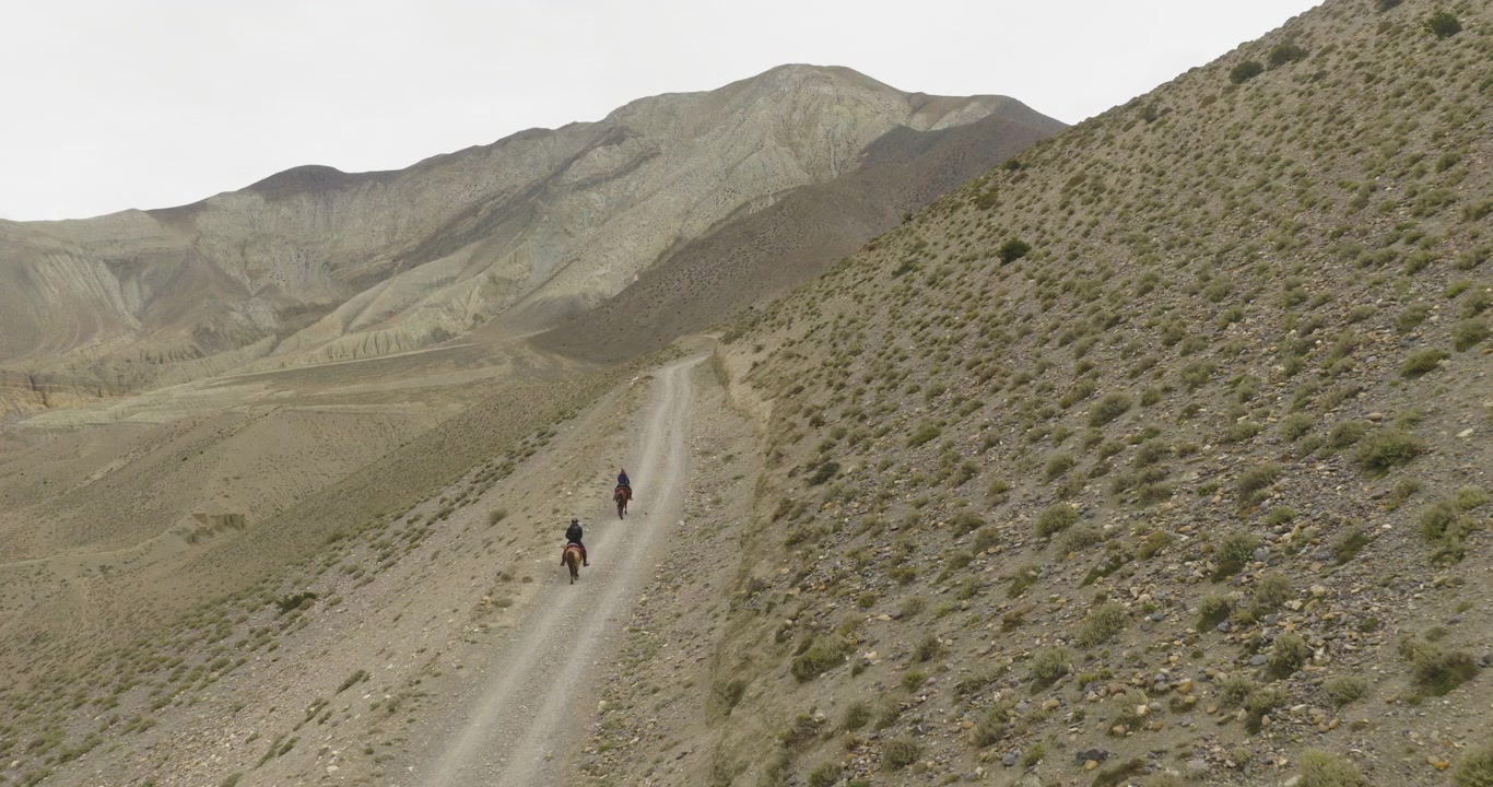 Mustang Nepal, aerial pull-out shot of a group of horseback riders navigating a steep sandy trail surrounded by arid slopes and a distant desert mountain peak.
