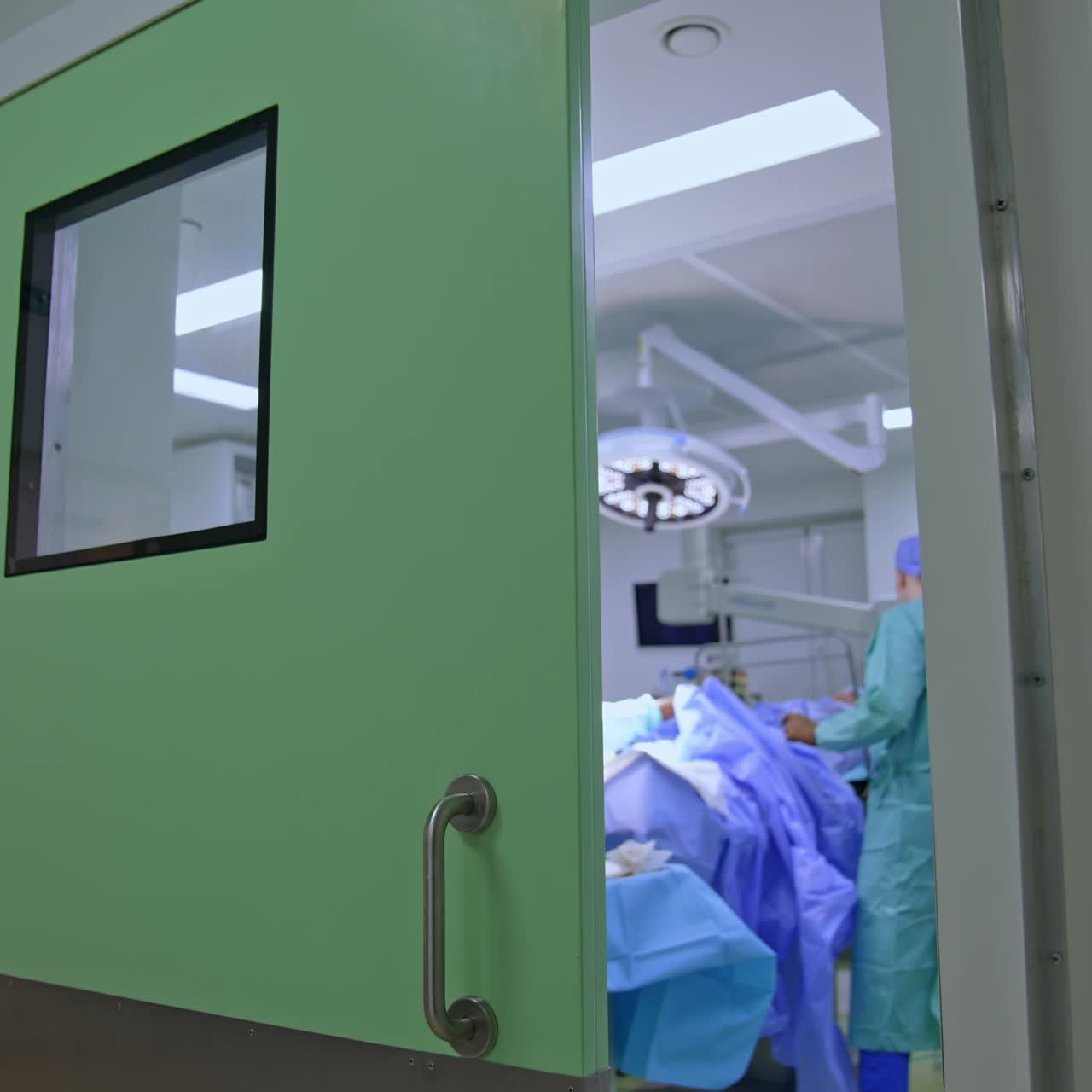 Puerta entreabierta que da a un quirófano moderno. Los médicos retiran las sábanas al paciente. El cirujano sale de la sala quitándose los guantes.