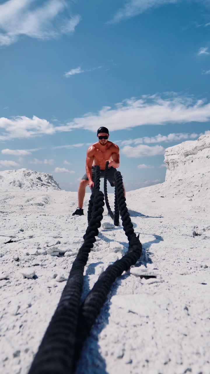 Bodybuilder trains his muscles with cable on the hill under blue sky. Shirtless sportsman wearing cap and sunglasses doing workout with battle ropes in sunny summer day outdoors. Vertical video