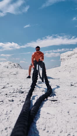 bodybuilder trains his muscles with cable on the hill under blue sky. shirtless sportsman wearing cap and sunglasses doing workout with battle ropes in sunny summer day outdoors. vertical video