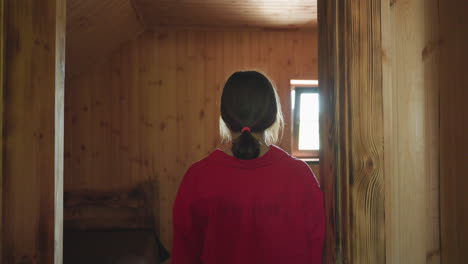 back view of young woman standing quietly by wooden door in cozy cabin room as soft window light glows ahead, creating peaceful mood in calm rustic setting with wood panel walls and warm tones