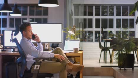 Tired-biracial-businessman-at-desk-with-computers,-talking-on-phone-in-office-at-night,-slow-motion