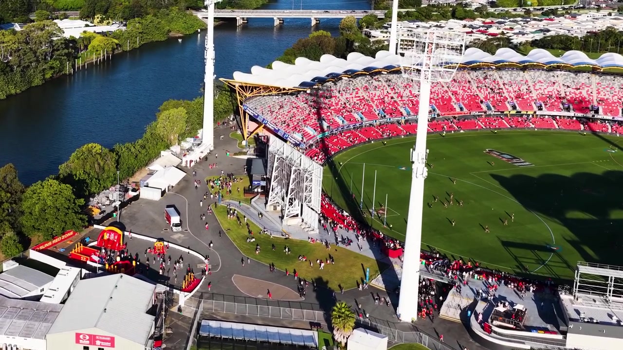 High-angle drone shot reveals a crowded sports arena, parking lot, and river in bright sunlight.