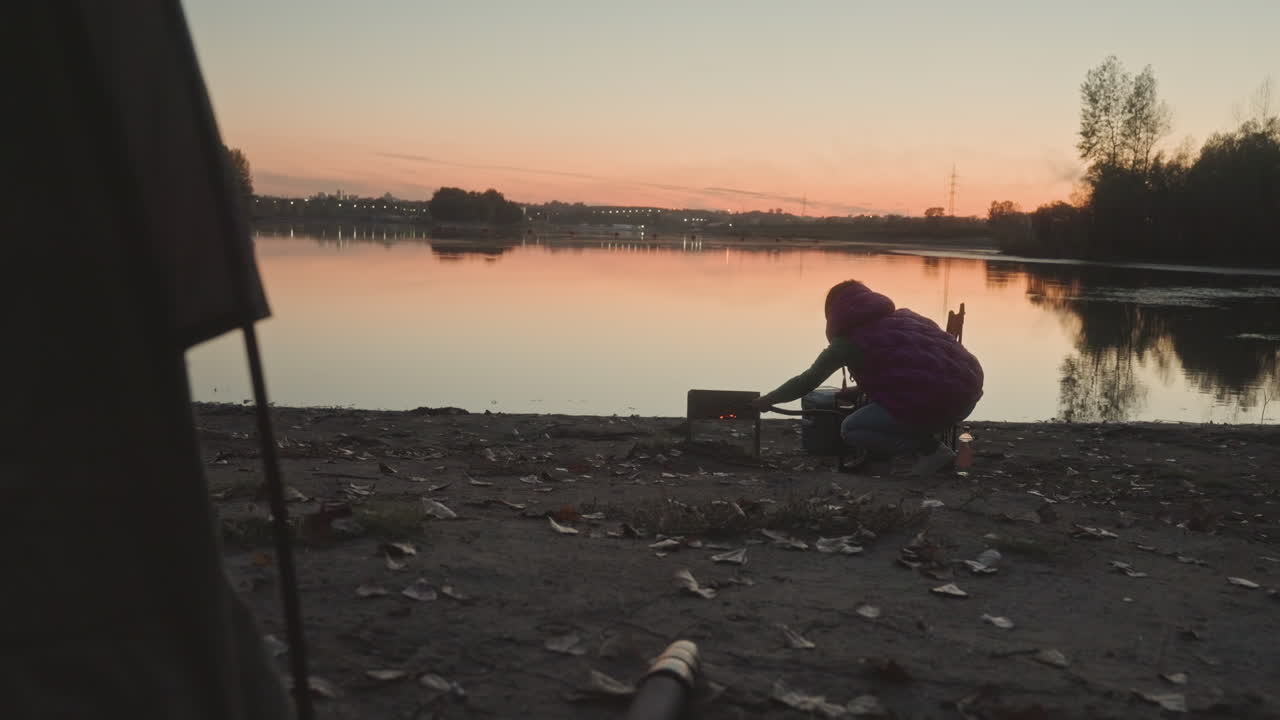 Sunset lake angler setting up fishing gear on rocky shore, twilight glow and water reflections, person crouched arranging rod and tackle boxes, portable stove and bucket nearby, calm breeze, focused