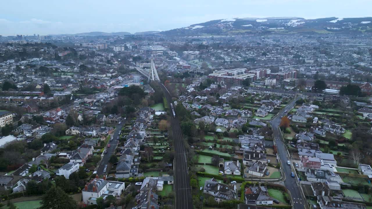 Aerial view of suburban South Dublin with train tracks, modern housing, and distant snowy mountains. Winter urban landscape with residential rooftops and soft overcast skies.