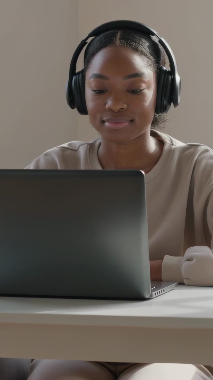 Vertical video: Sitting student wearing sweatshirt and headphones studying on laptop at table