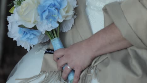 Bride-dressed-up-for-a-wedding-holding-a-bouquet-of-white,-blue-and-yellow-flowers-while-sitting-on-a-chair