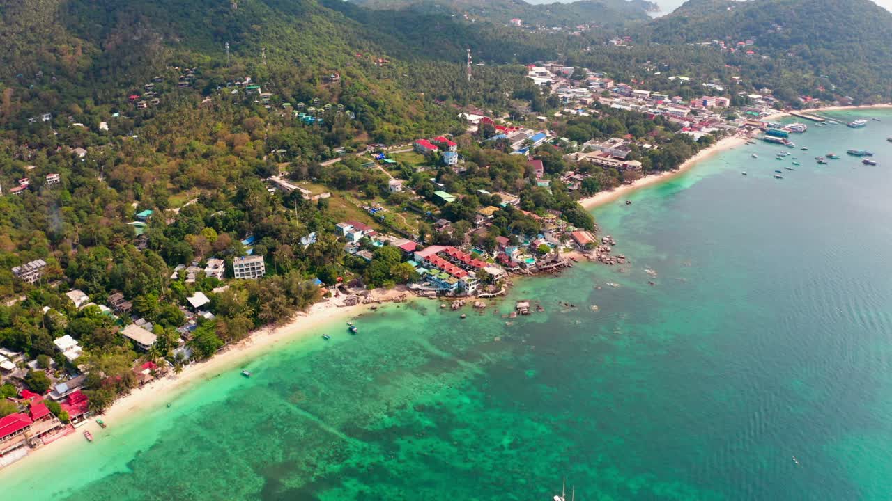 A very high aerial view from Koh Tao’s beach resort, showcasing its emerald waters, lush green hills, and boats floating in the bay.