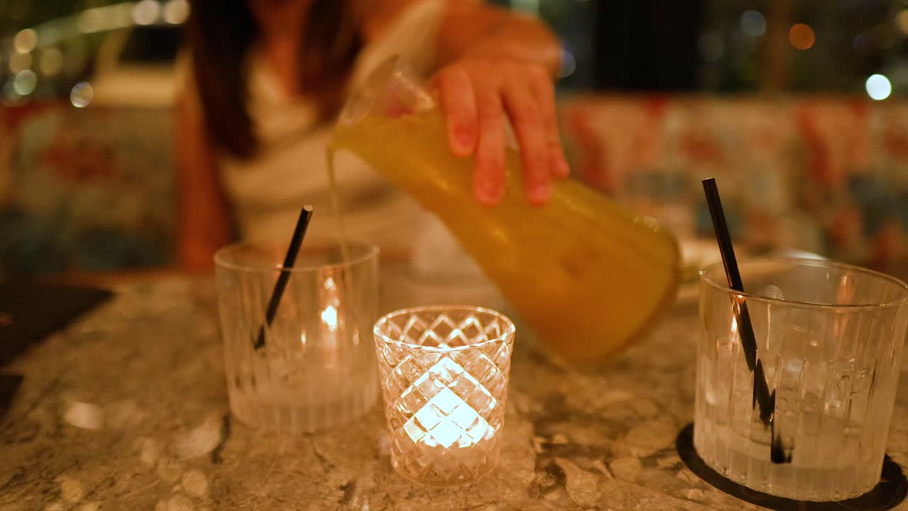 A person pours a yellow cocktail into a glass on a marble table, illuminated by candlelight in a cozy bar