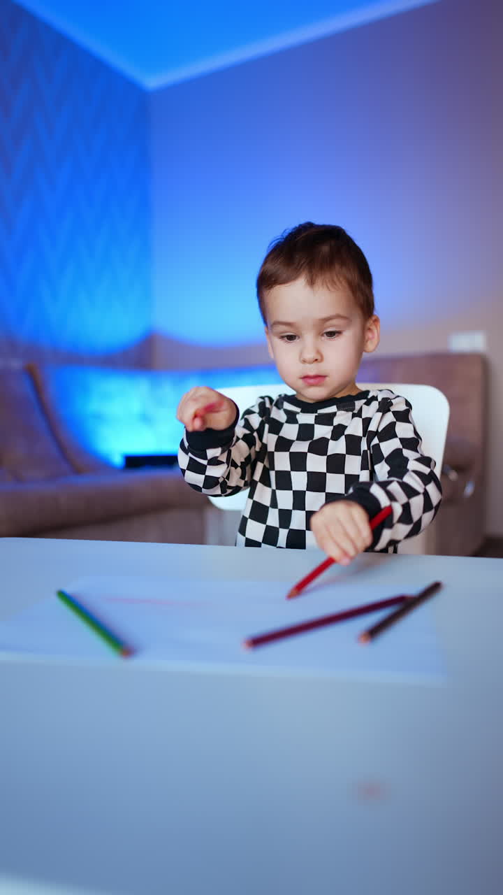 Caucasian kid in checkered shirt taps the pencils by the desk. Little boy starts to draw on the piece of paper. Vertical video.