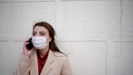 beautiful girl wearing protective medical mask and fashionable clothes speaks with a smart phone in front of wall