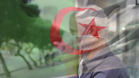 animation of flag of algeria waving over caucasian man wearing face mask in city street