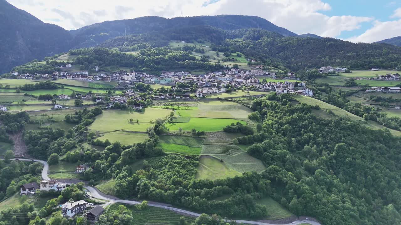 Aerial view of the Ponte Gardena village in the Italian Dolomites, with lush green fields, dense forests, winding roads, and majestic mountains