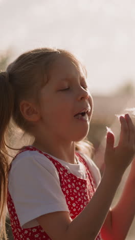 little girl puts piece of cotton candy into mouth and looks at sticky fingers against blurry fountain slow motion. spending time in amusement park
