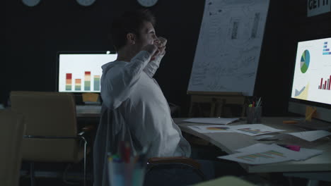 Tired-businessman-stretching-and-yawning-near-computer-in-night-office