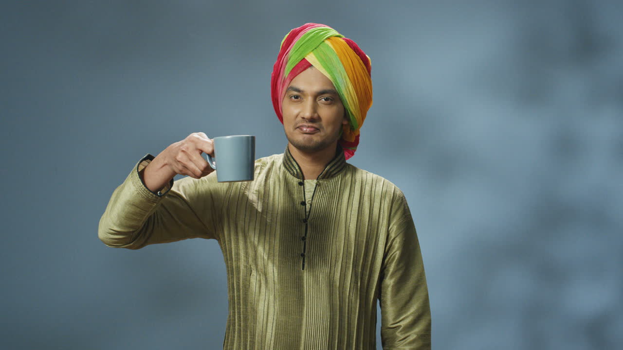 터번과 차 또는 커피를 마시고 카메라를 보고 웃는 전통 옷을 입은 젊은 행복한 인도 남자의 초상화