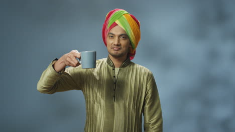 portrait of young happy indian man in turban and traditional clothes drinking tea or coffee and smiling at camera