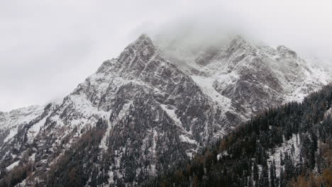 Aerial-cinematic-mountain-scape-drone-pan-to-the-left-view-of-colorful-trees-in-the-valley-and-snowy-mountains-in-background-during-cloudy-day-late-autumn-first-snow-in-Kauntertal,-Austria