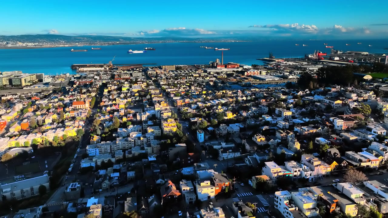 Tetti colorati di Dogpatch e Potrero Hill verso Oakland attraverso la baia. Ampia vista aerea di un reticolo di case multicolori, lungomare industriale, nave da carico su acque di un blu intenso e le colline dell'East Bay.