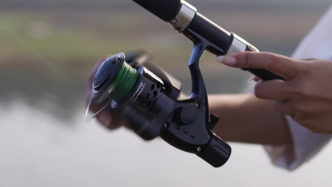 Vista ravvicinata di un mulinello e una canna da pesca tenuti dalle mani di una donna, che evidenzia la ricreazione all'aperto, la concentrazione e la pesca tranquilla al lago in pieno giorno.