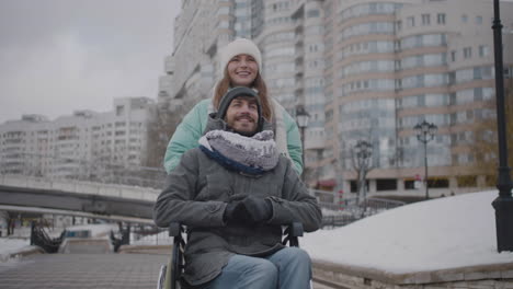 Happy-Caucasian-woman-taking-her-disabled-friend-in-wheelchair-for-a-walk-in-the-city-and-talking-together