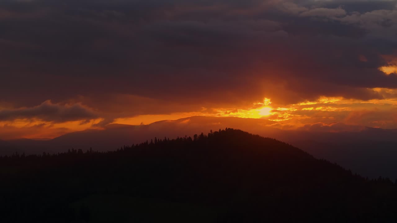 fotografía aérea de primer plano que muestra una puesta de sol naranja mientras arroja un brillo cálido entre un cielo nublado y una cresta montañosa con silueta, de alto contraste