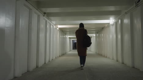 a back shot of a girl wearing a brown coat, jeans, white shoes, and a backpack as she walks slowly and faintly through a long, dimly lit underpass or tunnel