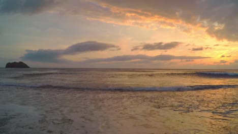 Una-Hermosa-Puesta-De-Sol-En-Una-Playa-Griega,-Con-Olas-Tranquilas-Y-Ondulantes-Y-Una-Formación-Rocosa-En-El-Océano