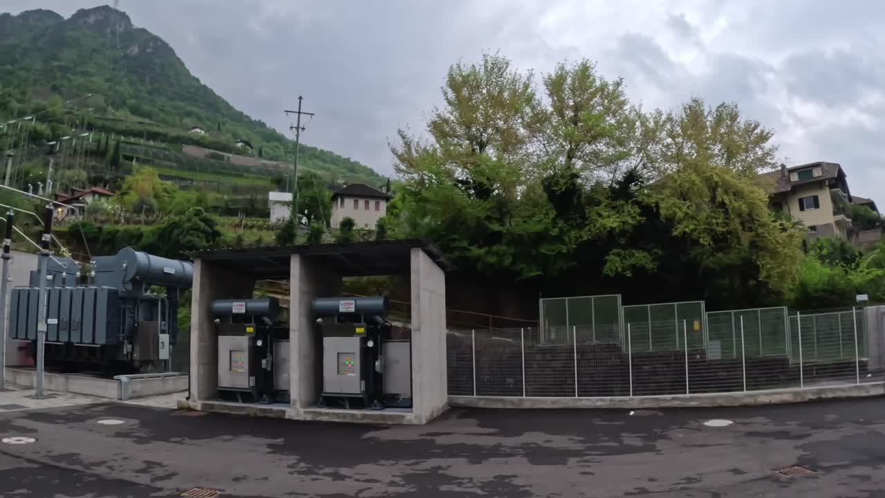 Camera pans substation yard showing large transformers, circuit breakers, and metal switchgear enclosures, set against the terraced Mendola foothills above Bolzano’s Talvera riverbank, slow motion