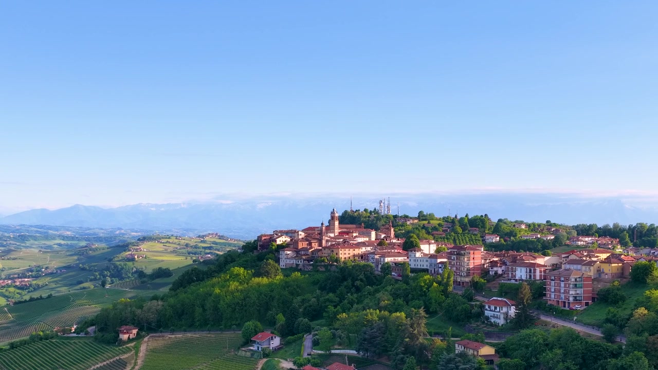Ampia ripresa aerea con drone di un villaggio storico immerso tra dolci colline verdi di vigneti.