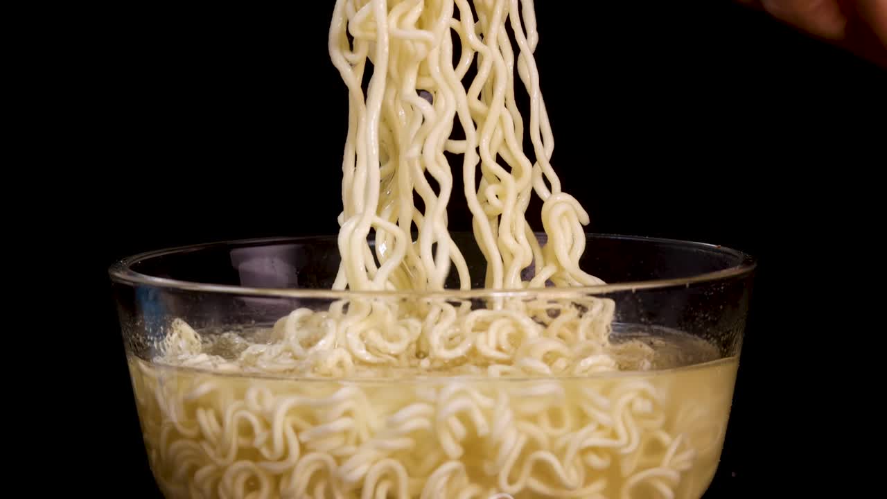 A hand skillfully lifts cooked instant noodles from a glass bowl using chopsticks, set against a black background with even, studio lighting and steady camera