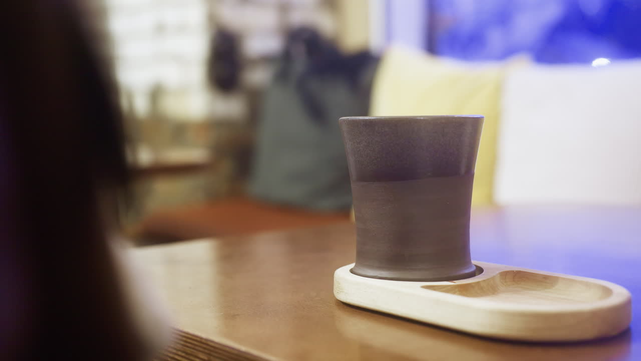 Side shot of lady's hand in brown shearling jacket gently lifting cup of coffee at cozy cafe table, with soft lighting enhancing the warm and inviting atmosphere perfect for relaxation