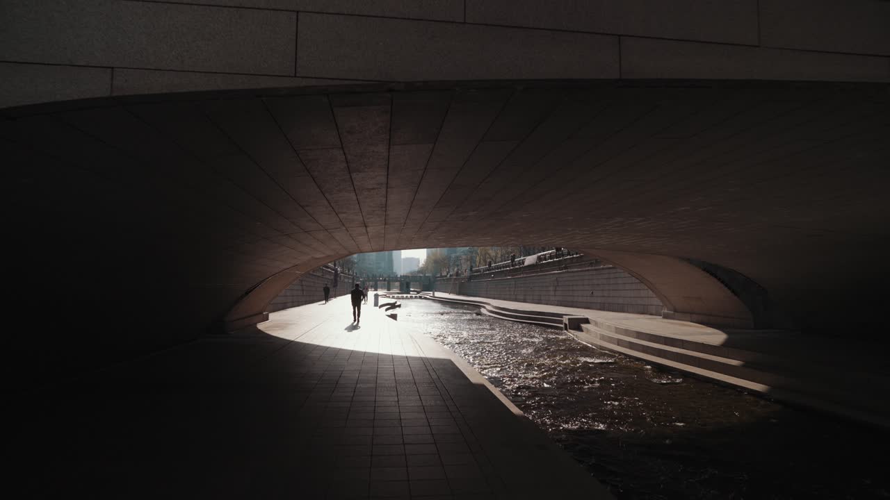 Person Walking Under a Bridge Along a City River in Sunlight
