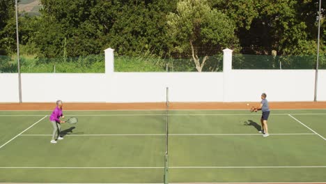 Video-of-happy-biracial-senior-couple-playing-tennis-on-tennis-court