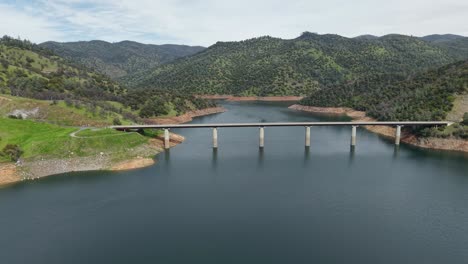 Captured from above, this aerial drone view showcases the Don Pedro Reservoir Bridge gracefully extending over the shimmering waters, framed by the surrounding golden hills and open skies.
