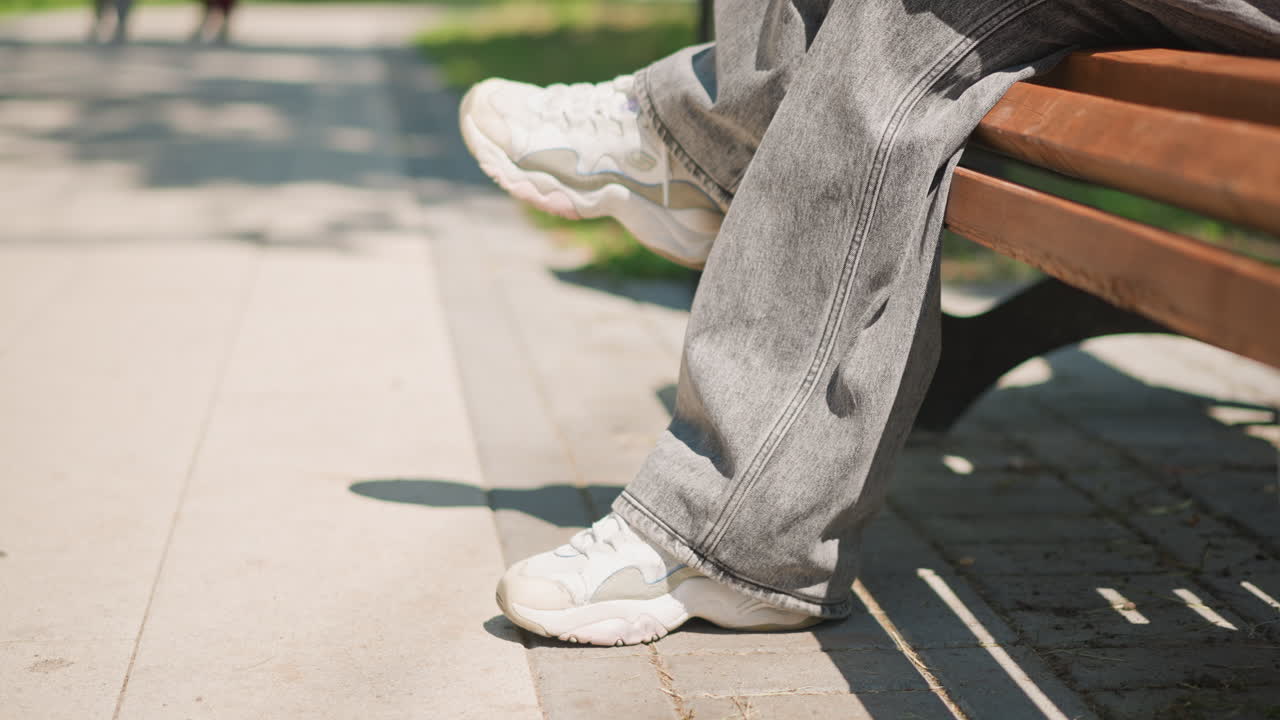 나무 벤치 아래 흰색 스니커즈, 좌석에 회색 트레이닝 바지, 햇볕이 드는 인도와 부드러운 그림자, 조용한 한낮의 휴식을 암시하는 신발 끈과 질감 있는 천의 클로즈업, 통근자 또는 학생