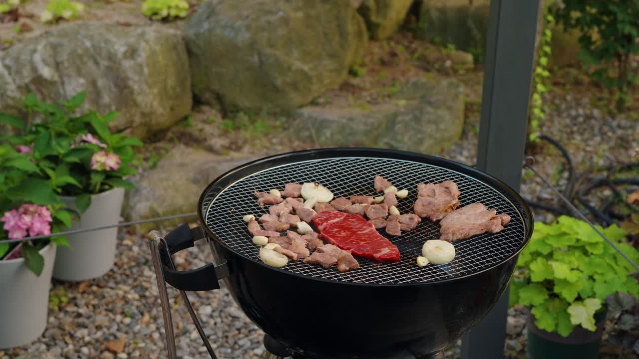 A woman seasons a variety of meats, including a fresh beef steak and pork, with a salt grinder while cooking on a charcoal barbecue during an outdoor garden party