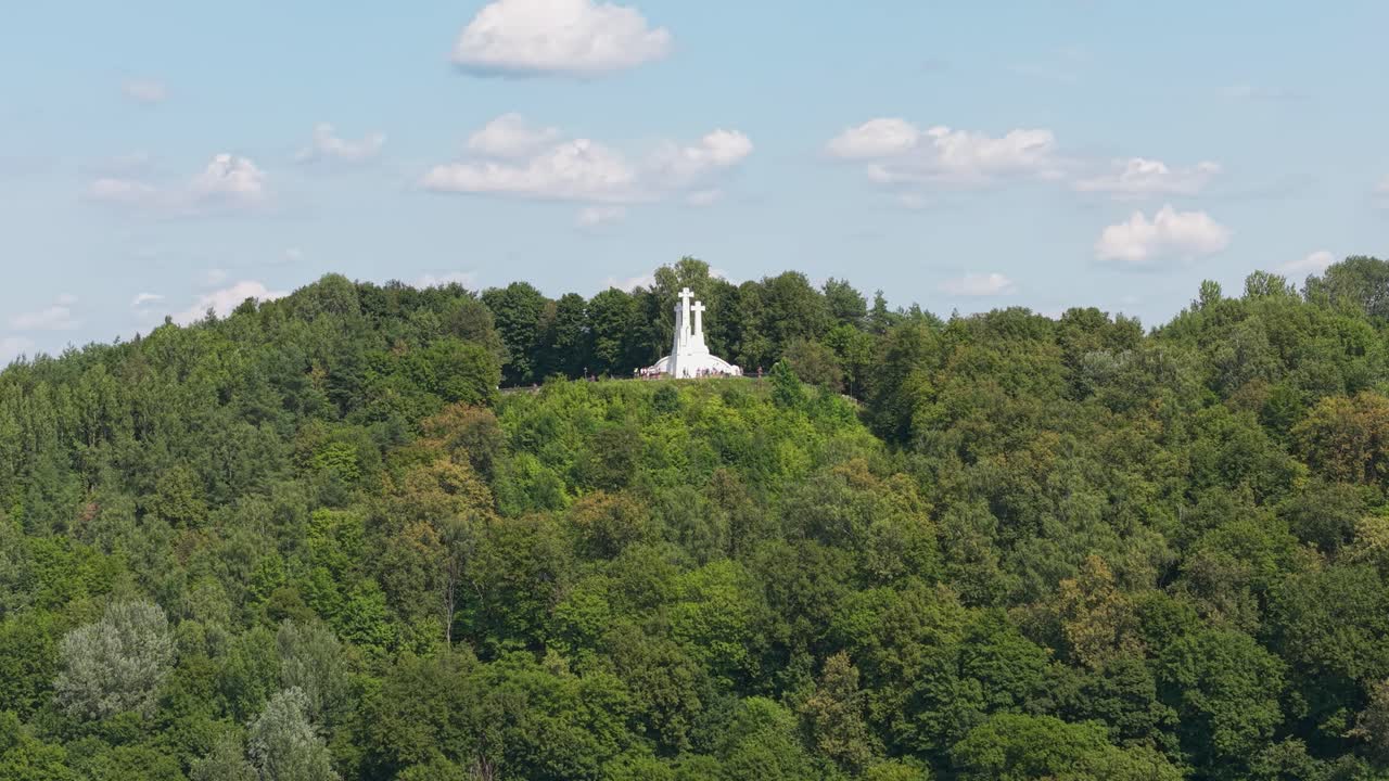 Aerial view of the iconic Three Crosses monument standing atop a lush green hill in Vilnius, Lithuania, surrounded by dense forest under a bright blue summer sky