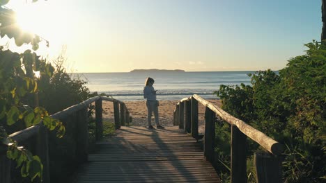 Woman-stands-on-wooden-deck-by-the-beach-at-sunrise-golden-hour-using-smartphone-with-running-suit-and-sneakers