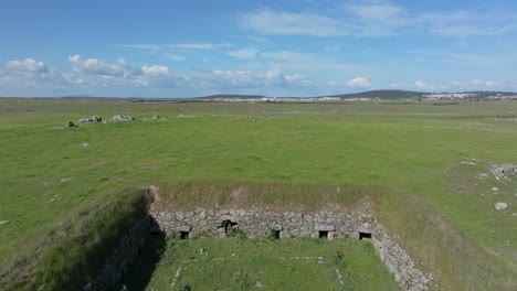ascending and reversing drone flight inside an 18th-century stone structure used for pig farming. multiple side entrances with lintels visible. grass growing on the roof, surrounded by green pastures.