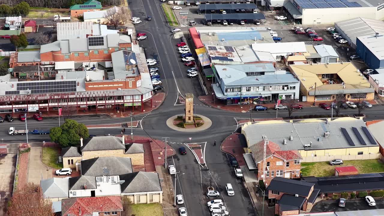 Aerial footage moves above a small town center, passing a clock tower roundabout, commercial buildings, and residential streets in soft daylight