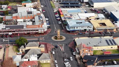 aerial footage moves above a small town center, passing a clock tower roundabout, commercial buildings, and residential streets in soft daylight