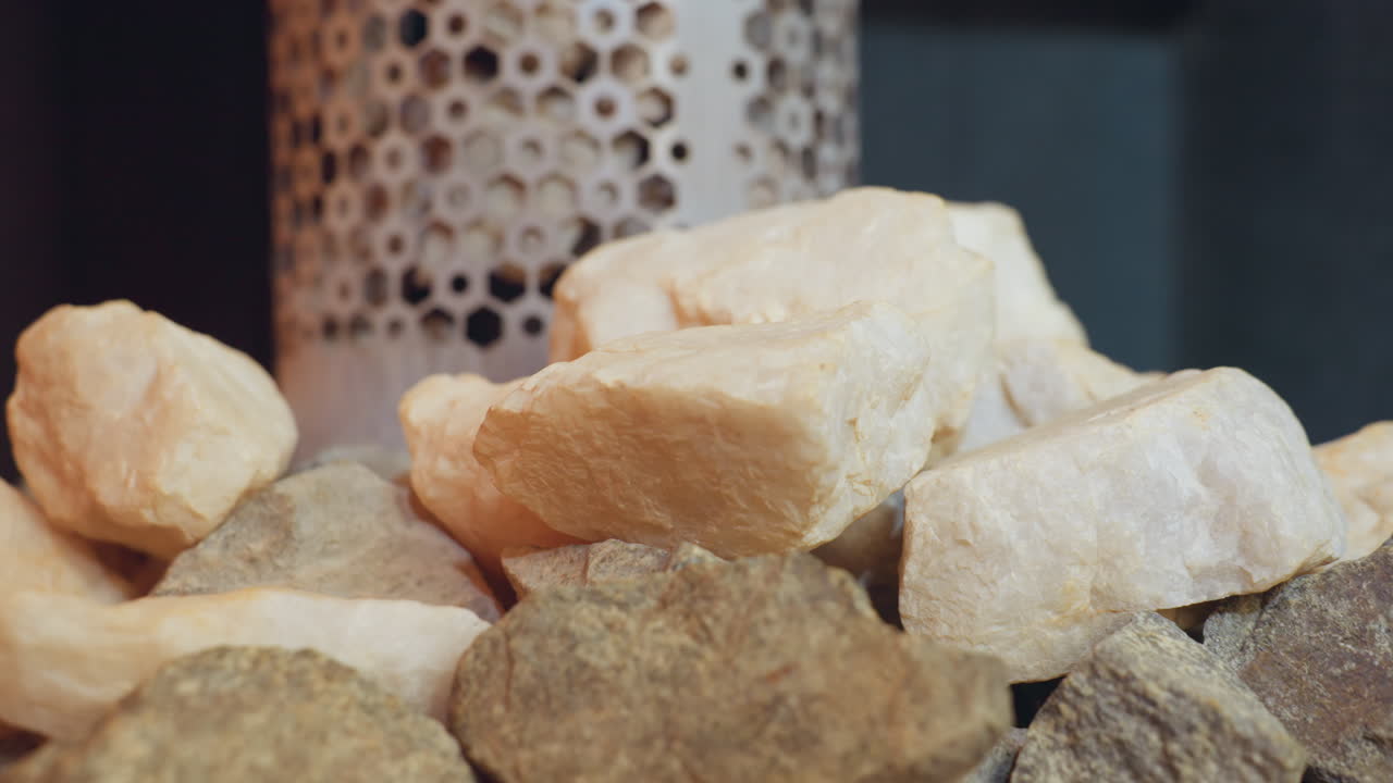 Close up of rough sauna stones stacked in front of perforated metal heater inside steam room, showing light and dark textures of rocks used for generating heat in wellness and relaxation environments