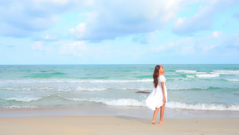 Una-Joven-Atractiva-Con-Un-Vestido-Blanco-Camina-Lentamente-Por-Una-Playa-De-Arena-Hacia-El-Oleaje