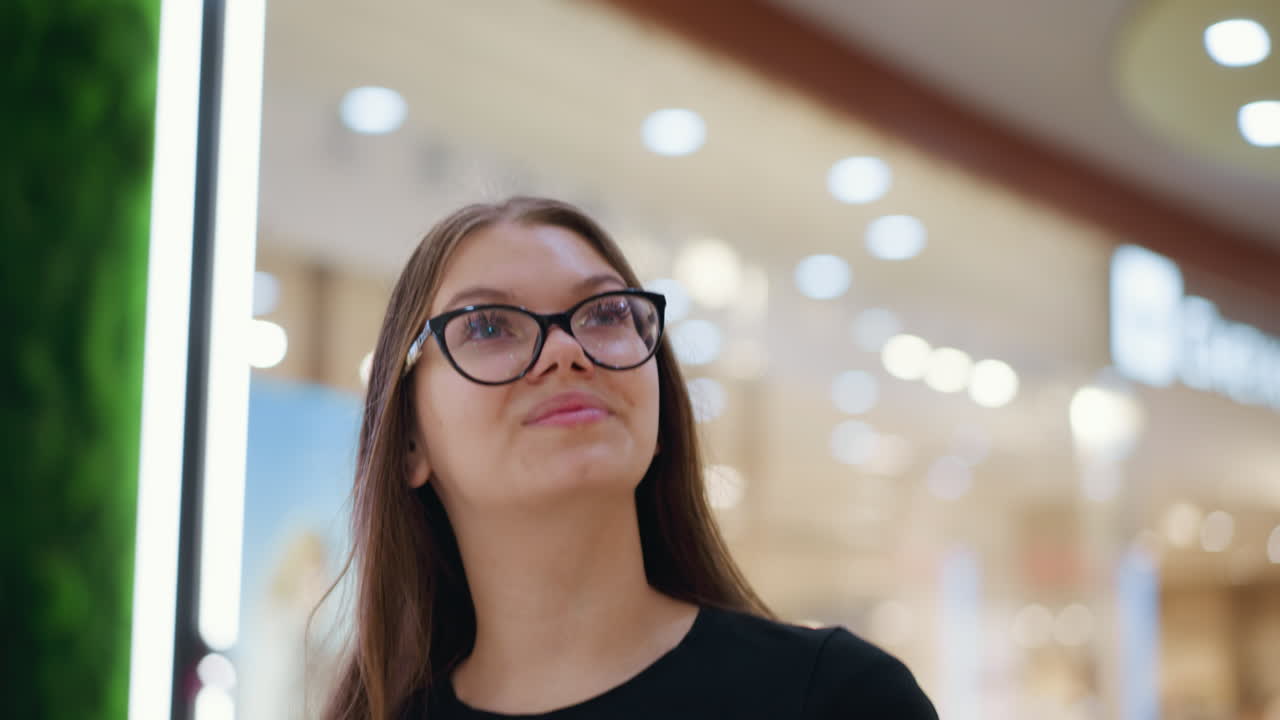 mujer joven con gafas caminando en un centro comercial con luz bokeh en el fondo, ambiente de compras casual, fondo borroso con enfoque en el comprador, entorno moderno del centro comercial