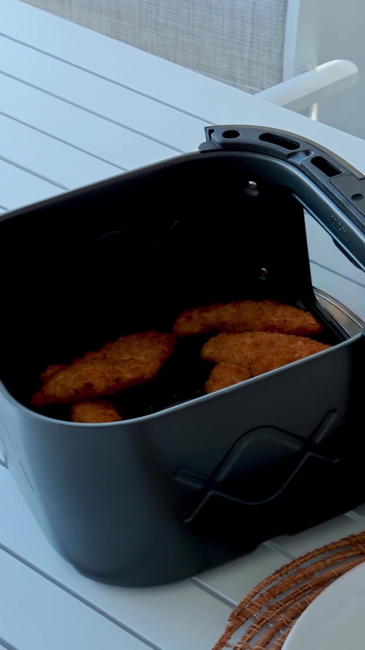En person åbner en airfryer, tager kurven med panerede koteletter ud og placerer den på et lyst, moderne spisebord. Naturligt dagslys, stabilt kamera.